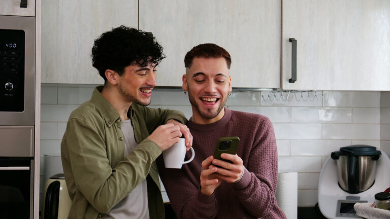 Zwei junge Männer stehen in einer Küche und schauen gemeinsam auf ein Smartphone.