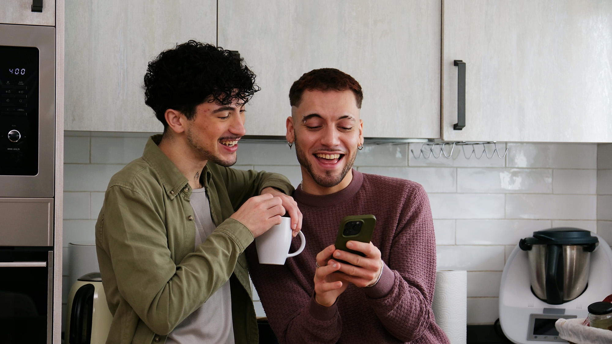 Zwei junge Männer stehen in einer Küche und schauen gemeinsam auf ein Smartphone.