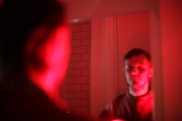Mann blickt im Badezimmer in den Spiegel unter rotem Licht – Symbolbild für Stress und Belastung in der Angehörigenpflege