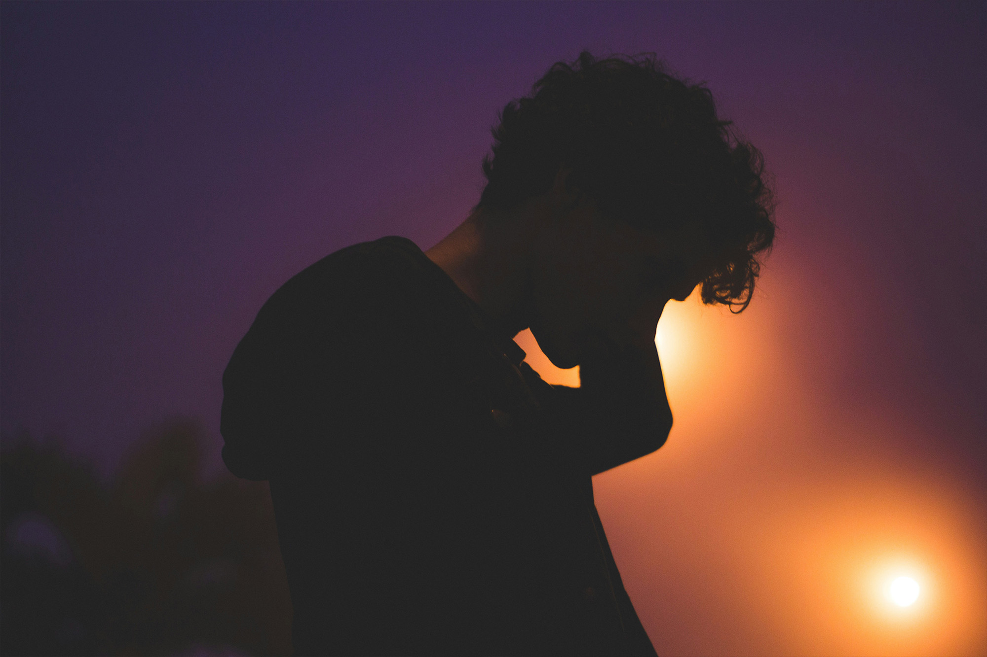 Dunkle Silhouette eines Menschen mit gesenktem Kopf und einer Hand an der Stirn vor einem orange-violetten Abendhimmel.