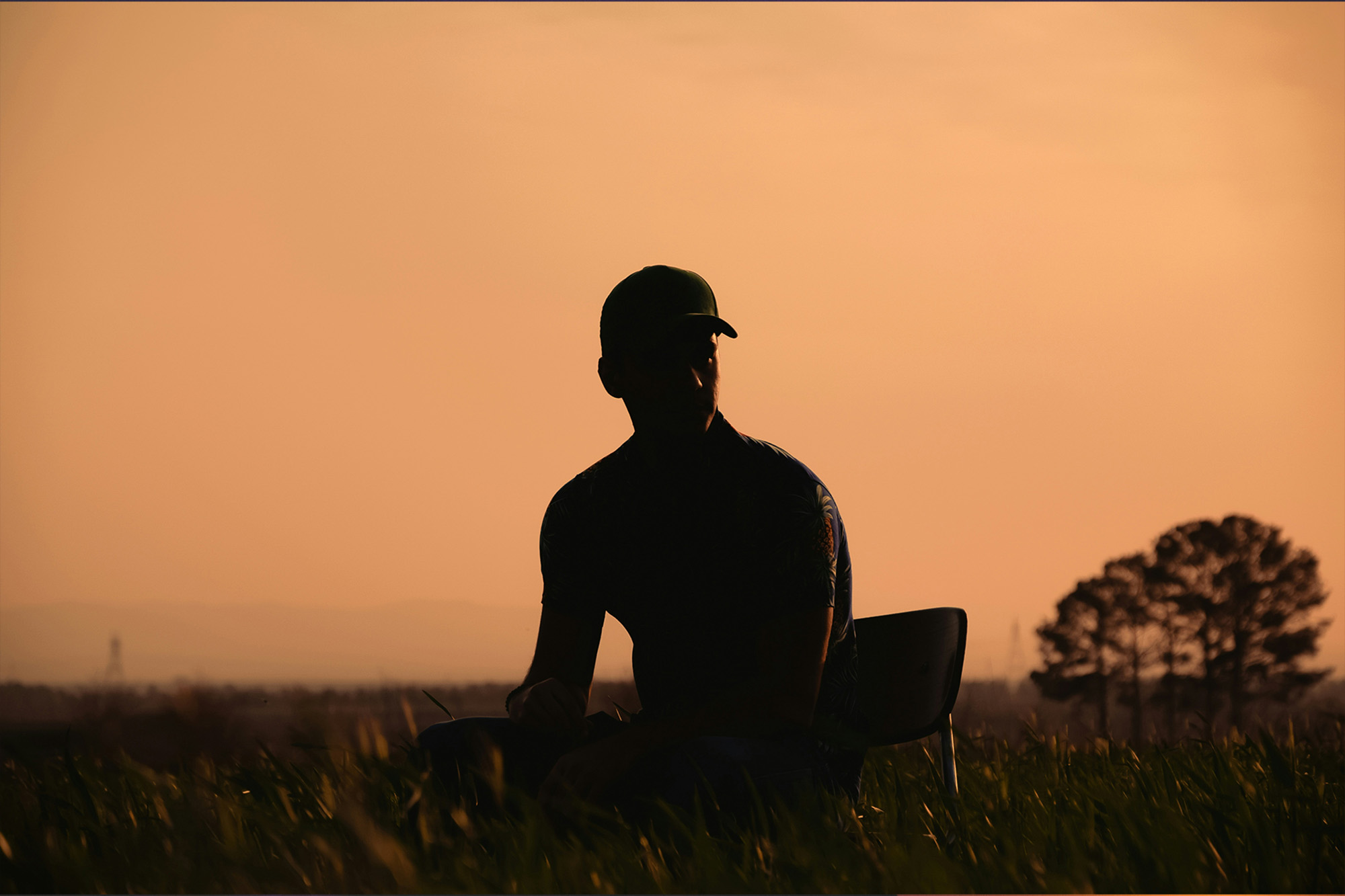 Silhouette eines sitzenden Menschen in einer Wiese bei Sonnenuntergang vor einem orangefarbenen Himmel.